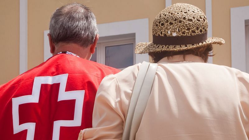 Dues persones caminen juntes a l'exterior, una amb un armilla vermella de la Creu Roja i l'altra amb barret de palla, a prop d'un edifici de façana ocre.