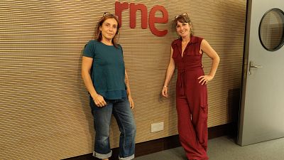 Dos mujeres posan de pie en un estudio de radio con paneles ac�sticos de madera y el logotipo rojo de rne en la pared central.
