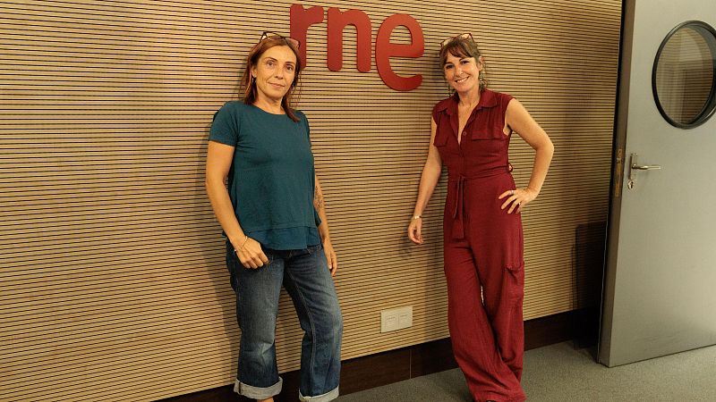 Dos mujeres posan de pie en un estudio de radio con paneles acústicos de madera y el logotipo rojo de rne en la pared central.