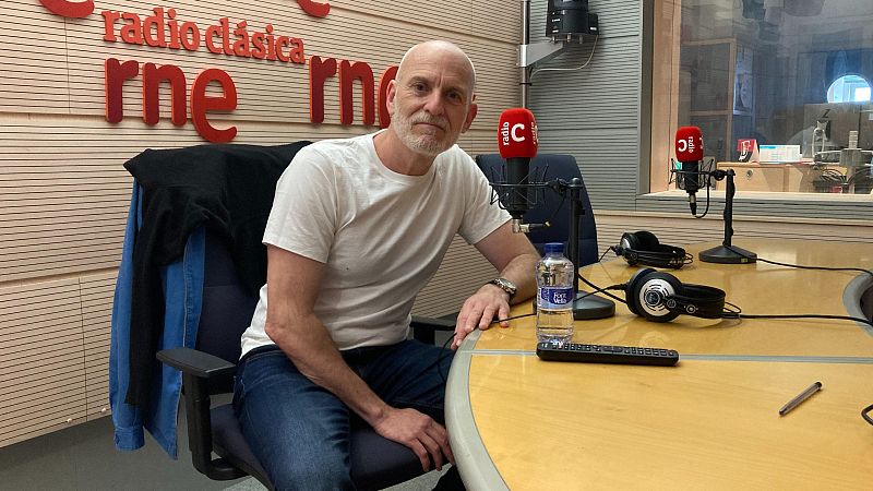 Un hombre con barba y camiseta blanca sentado en un estudio de radio con micr�fonos y el logotipo de Radio Cl�sica al fondo.