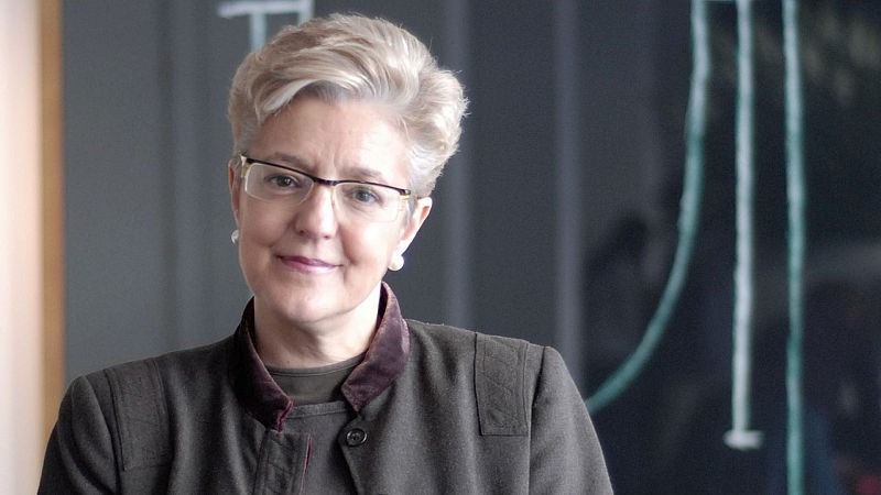 Plano medio de la escritora Anna Caball� sonriendo levemente, con cabello corto rubio, gafas y abrigo verde oliva ante un fondo de pizarra.