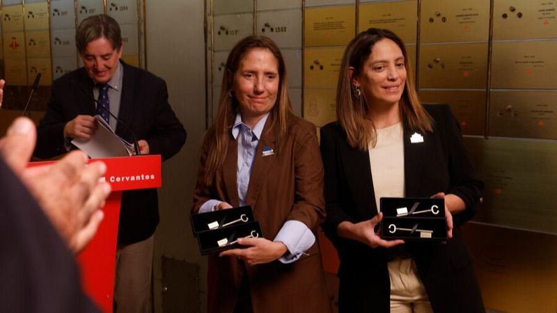 Dos mujeres posan sonrientes sosteniendo llaves ceremoniales en una cámara acorazada, flanqueadas por taquillas metálicas y un hombre leyendo junto a un atril.