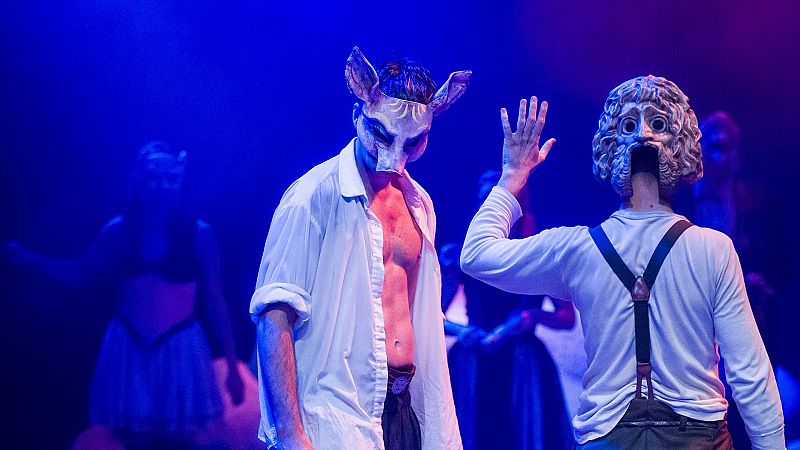 Escena teatral iluminada en azul con dos actores en primer plano usando m�scaras mitol�gicas.