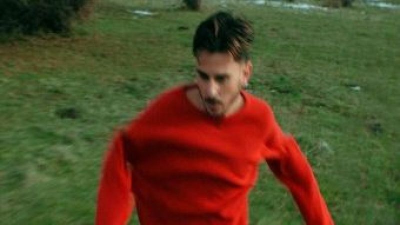 Un joven con jersey rojo de punto y pendiente corre por una pradera verde bajo una luz diurna.