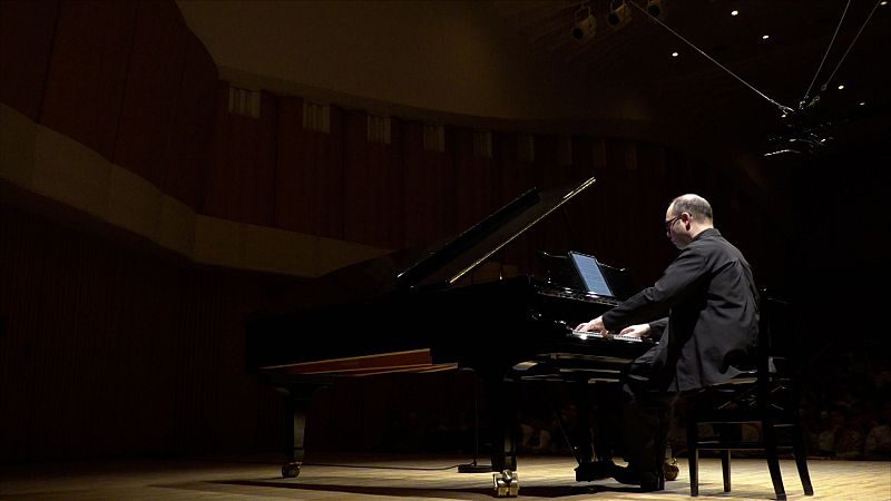 El pianista Alexander Melnikov toca un gran piano de cola negro con una tableta digital en el atril bajo una iluminaci�n c�lida en un auditorio.