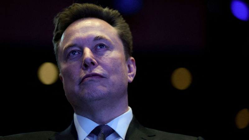 Primer plano contrapicado de Elon Musk con traje formal y expresi�n seria sobre un fondo teatral con luces desenfocadas.