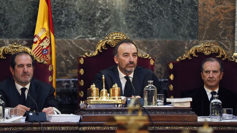 El magistrado Manuel Marchena habla ante un micr�fono en el estrado del Tribunal Supremo, acompa�ado por otros jueces y con la bandera de Espa�a al fondo.