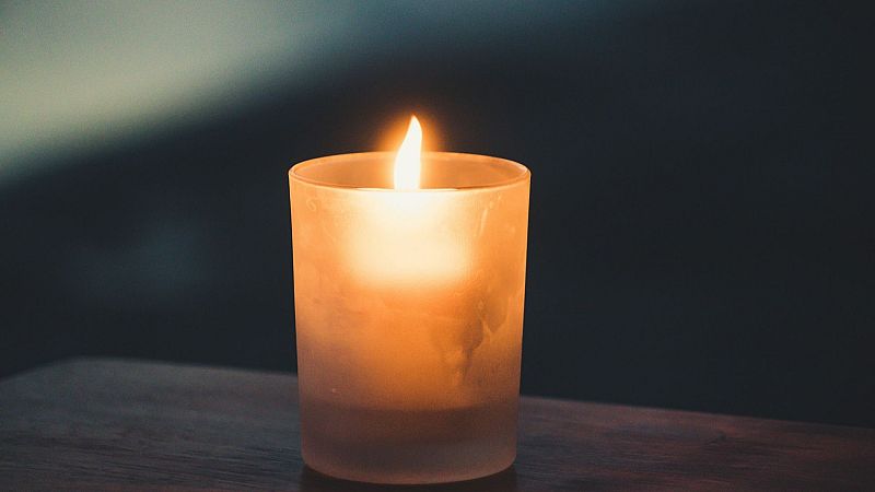 Vela encendida en un vaso de vidrio mate sobre madera oscura, con una llama c�lida que destaca sobre un fondo azulado en penumbra.