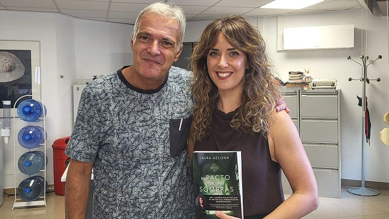 La escritora Laura Azcona sostiene su novela «El pacto de las sombras» sonriendo junto a un compañero en una oficina.