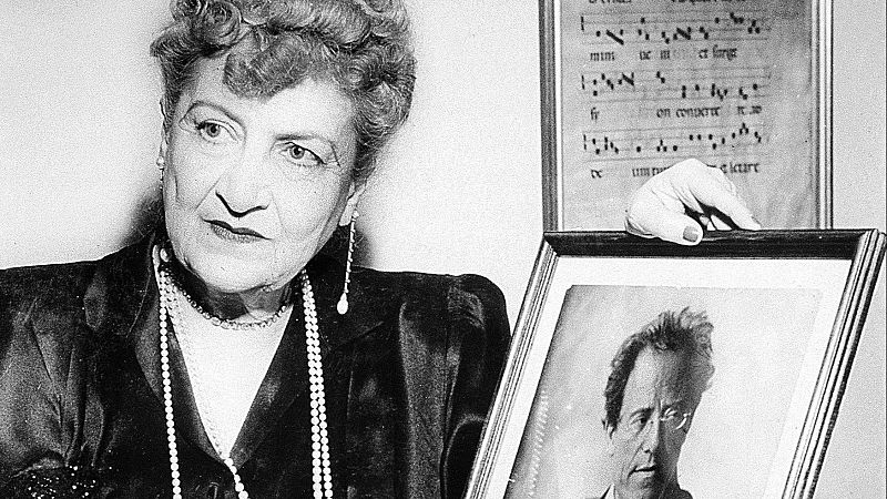 Alma Mahler de anciana, con pelo rizado, elegante ropa oscura y collares de perlas, sostiene un retrato enmarcado de Gustav Mahler.