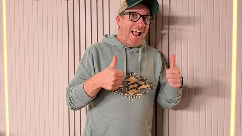 Hombre sonriente con sudadera verde, gorra y gafas hace gestos de aprobaci�n en un estudio de grabaci�n con paneles de madera y luces LED.