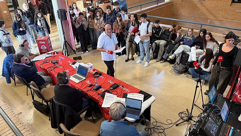 Emisión radiofónica en directo con periodistas sentados en una mesa corporativa roja, mesa de mezclas y ordenadores.
