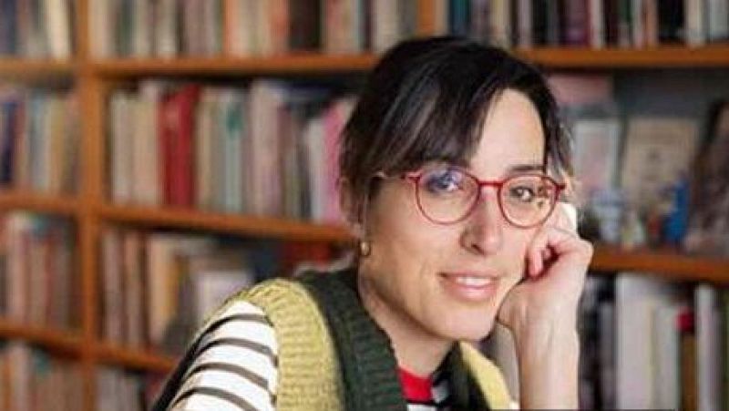 La periodista Carlota Visiers sonríe suavemente a la cámara en una biblioteca, luciendo gafas rojas, chaleco mostaza y camiseta de rayas.