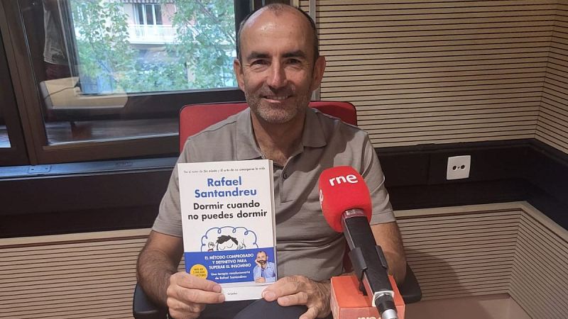 El escritor Rafael Santandreu sostiene su libro en un estudio de radio con paneles de madera y un micrófono de RNE.