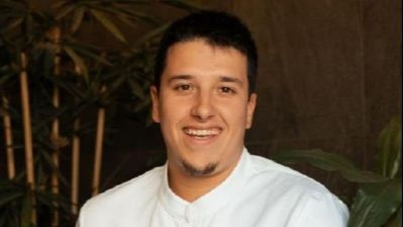 Plano medio corto del chef Aarón Ortiz sonriendo, con chaquetilla blanca y fondo vegetal oscuro.