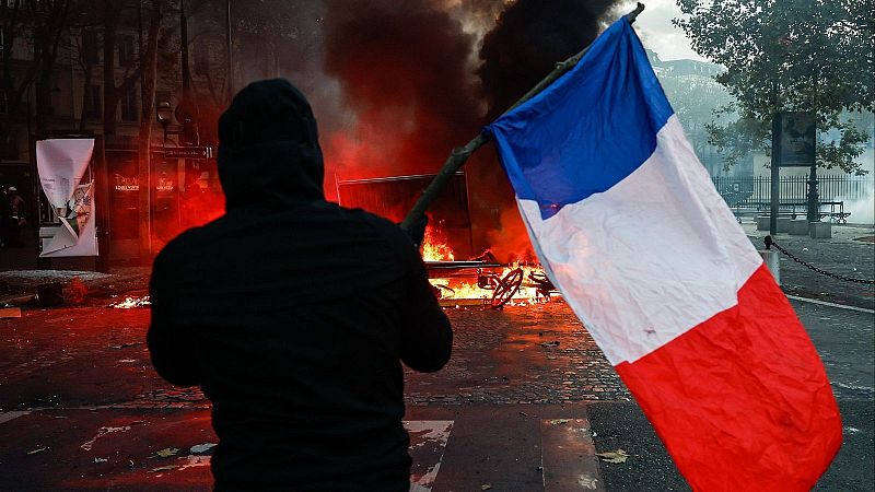 Manifestante de espaldas ondea la bandera de Francia frente a una barricada en llamas en una calle adoquinada.