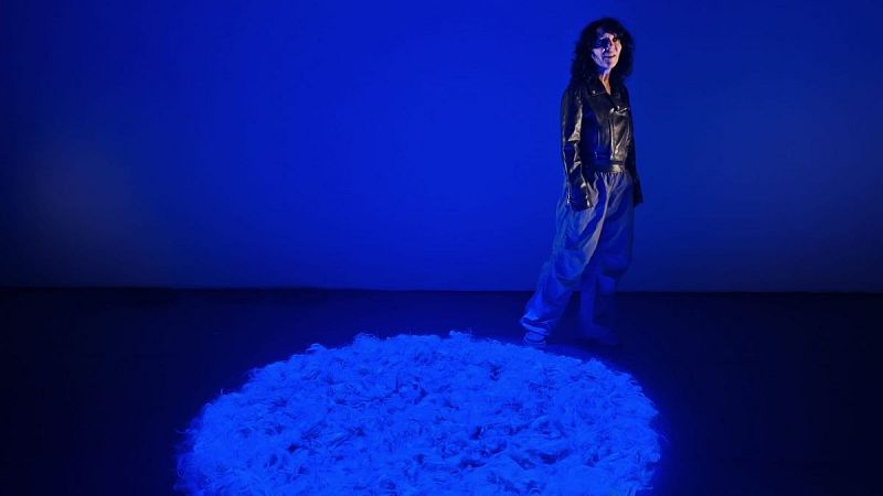 Instalación artística iluminada con luz azul eléctrico donde una mujer de cabello rizado y cazadora negra posa junto a una escultura circular texturizada.