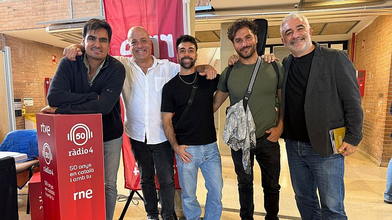 Cinco hombres posan sonrientes junto a un atril promocional rojo de R�dio 4 que celebra su 50 aniversario en un pasillo con paredes de ladrillo visto.