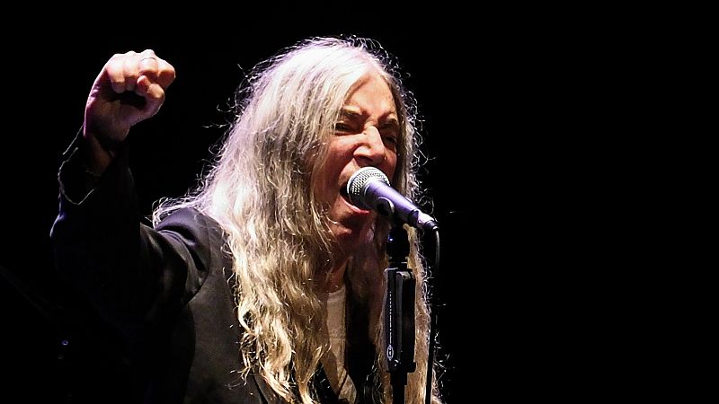 Patti Smith canta apasionadamente en directo sobre un escenario oscuro, con el puño derecho en alto y una intensa iluminación focal que resalta su melena plateada.