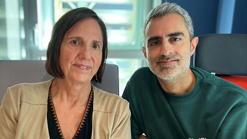 Dos personas sonríen juntas en un estudio de radio con ventanales al fondo; ella lleva chaqueta beige y él una sudadera verde.