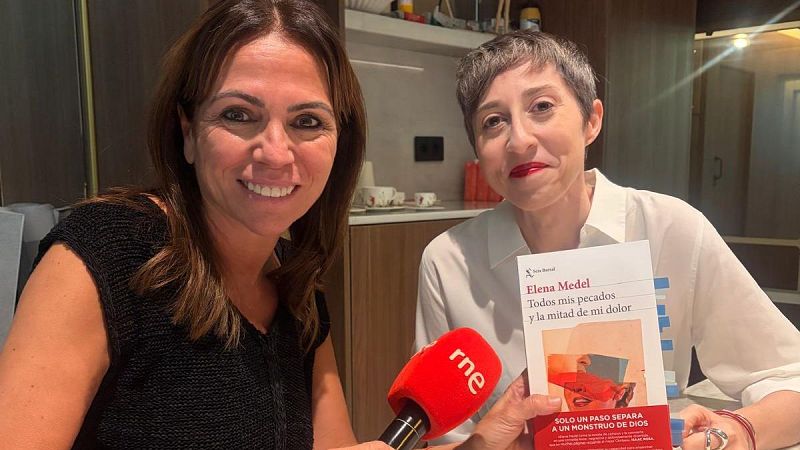 Dos mujeres sonrientes sostienen un libro durante una entrevista en un estudio de radio, sujetando un micrófono con espuma roja.