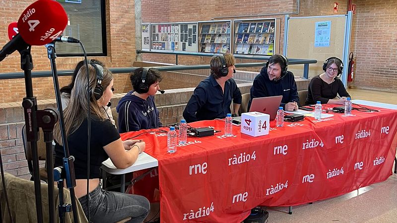 Cinco participantes con auriculares participan en una retransmisión de radio alrededor de una mesa en un estudio interior.
