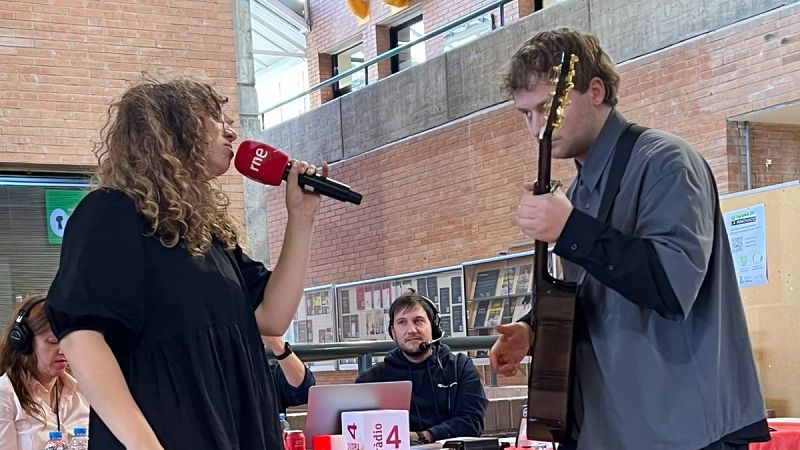 La cantante J�lia Sabat� interpreta una canci�n en directo con un micr�fono junto a un guitarrista en un estudio de radio con paredes de ladrillo y t�cnicos al fondo.