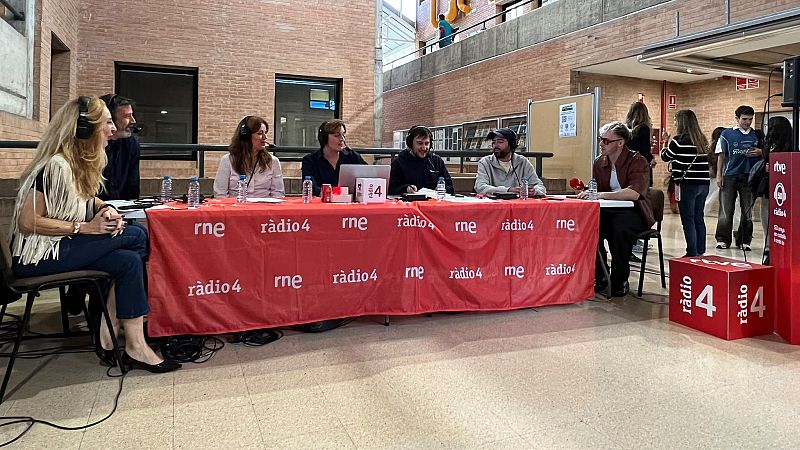 Emisi�n en directo de un programa de radio con siete personas sentadas ante una mesa con fald�n rojo y micr�fonos en un vest�bulo moderno.