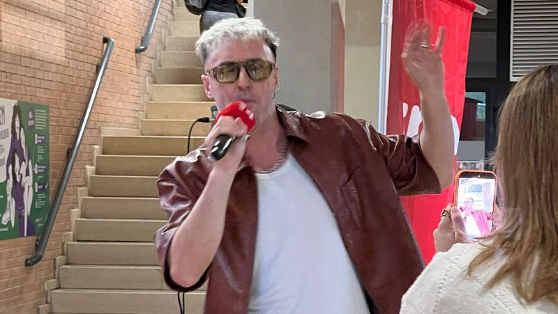 Plano medio de Markos amb K cantando con micrófono, gafas de sol y chaqueta de cuero marrón, mientras alguien le graba con un móvil en primer plano.