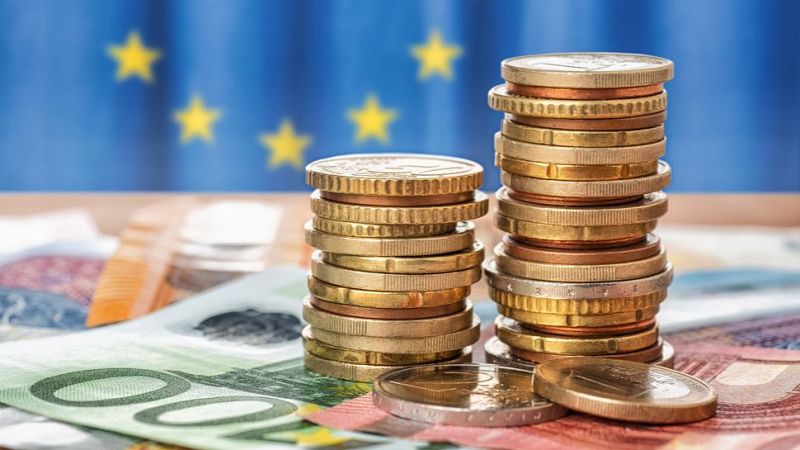 Primer plano de billetes y pilas de monedas de euro sobre una mesa, con la bandera de la Unión Europea desenfocada al fondo.