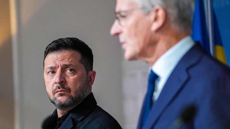 Plano medio de Volodímir Zelenski con gesto serio durante una rueda de prensa, con un dirigente desenfocado en primer término y la bandera de Ucrania al fondo.