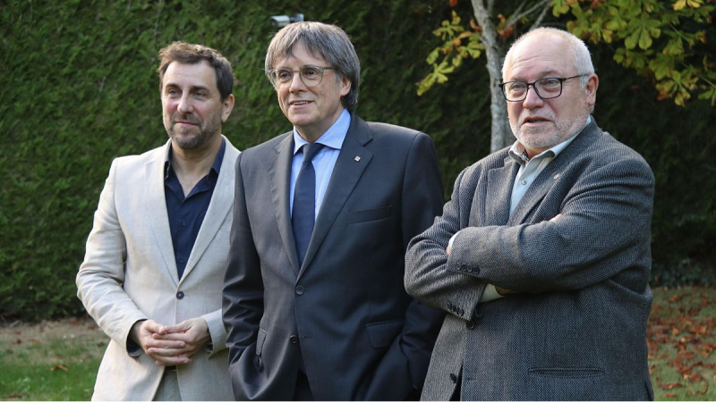 Tres hombres de pie en un exterior con seto de fondo; en el centro Carles Puigdemont con traje, flanqueado por Toni Com�n y Llu�s Puig.