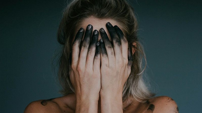Mujer de hombros descubiertos cubriendo su rostro con las manos manchadas de negro sobre un fondo gris.
