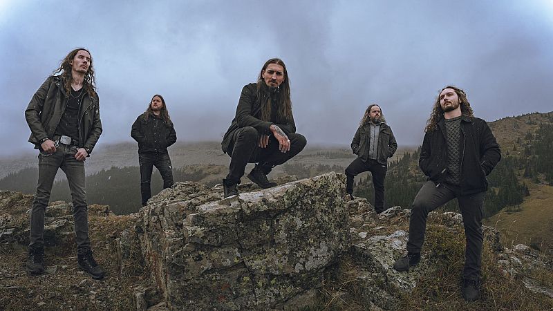 Cinco músicos de rock y metal posan con aspecto serio en una colina rocosa bajo un cielo nublado de alta montaña.