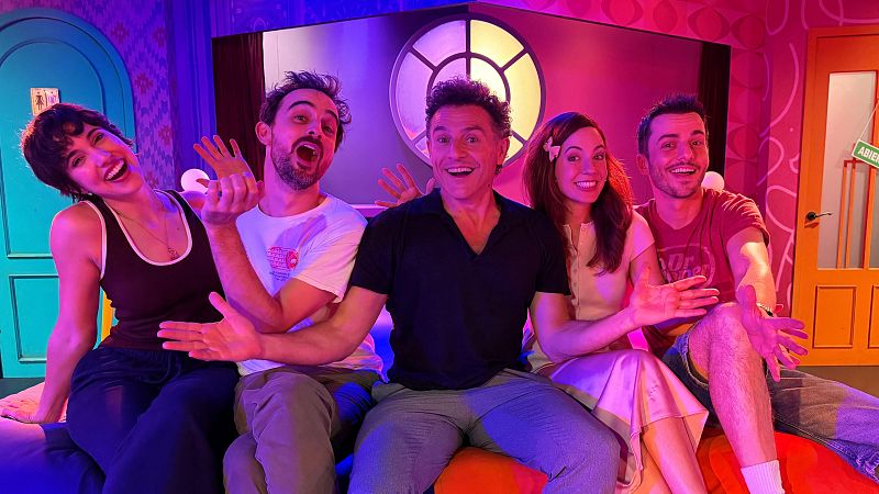 Cinco personas sonrientes posan juntas en una cama dentro de un colorido decorado teatral iluminado con luces violetas y azules, acompañando al periodista Miguel Rabaneda.