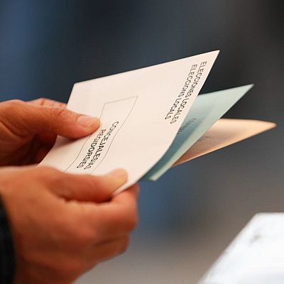 Persona sosteniendo sobres electorales blancos y de colores en una jornada de votación local, con urnas transparentes al fondo.
