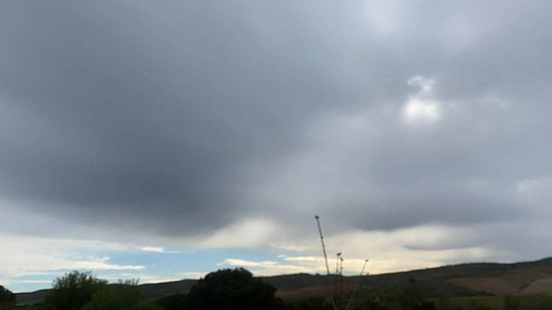 Paisaje rural bajo un cielo cubierto de oscuras nubes de tormenta, con colinas suaves y árboles a contraluz en primer plano.