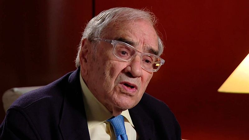 Primer plano del pol�tico Rodolfo Mart�n Villa con expresi�n seria, gafas transparentes, traje oscuro, camisa amarilla y corbata azul ante un fondo rojo.