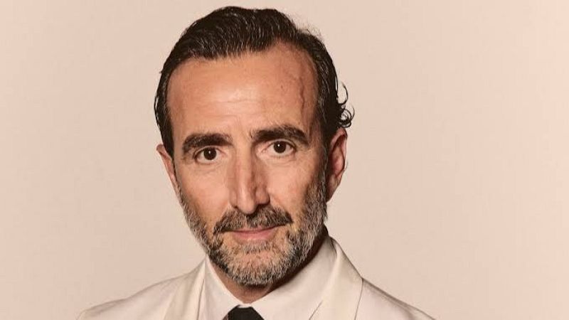 Primer plano del actor Enrique Arce con traje formal blanco y corbata negra sobre fondo beige claro.