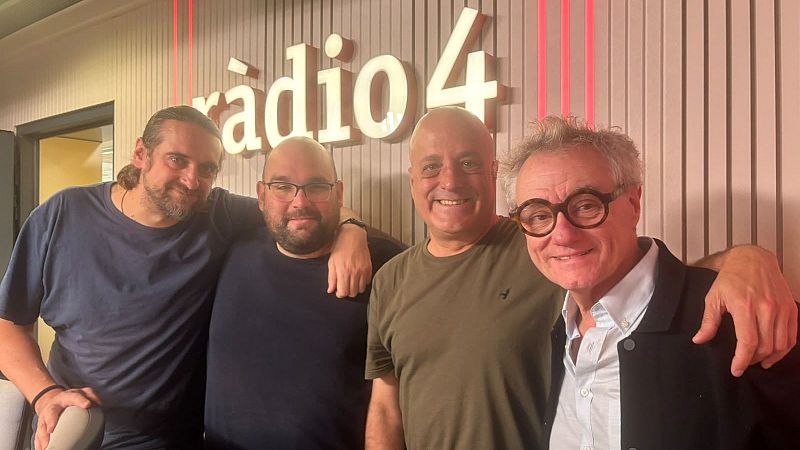 Cuatro hombres posan juntos y sonrientes en un estudio de radio con paneles de madera y el letrero iluminado.