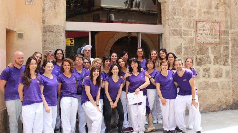 Grupo de profesionales sanitarios y sociosanitarios sonrientes posando uniformados frente a la entrada de piedra de un edificio con puerta acristalada.