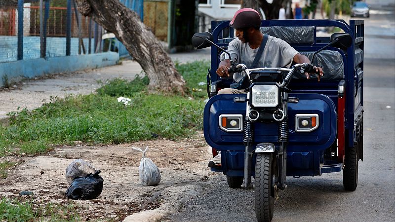 Un hombre con casco marrón conduce un motocarro azul por una calle residencial, pasando junto a bolsas de basura acumuladas en la acera.