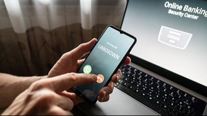 Primer plano de unas manos sosteniendo un m�vil con una llamada entrante desconocida junto a un ordenador con banca online.