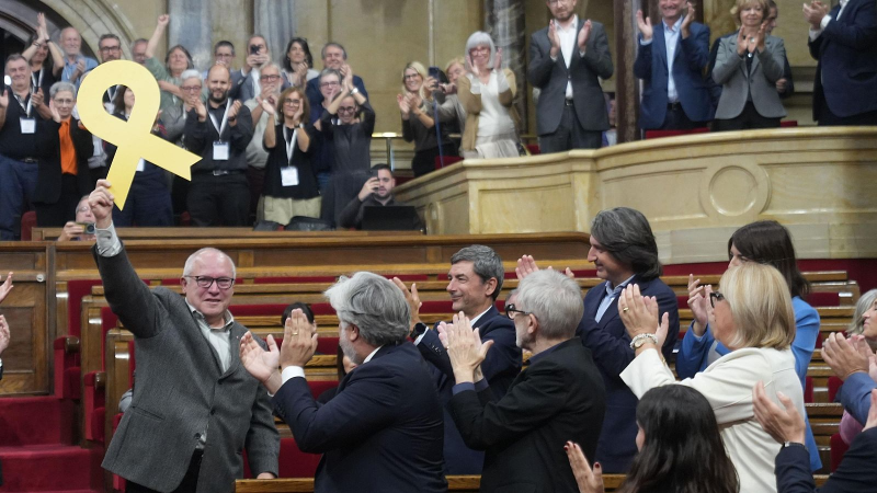Diputados catalanes aplauden de pie mientras un pol�tico sostiene un gran lazo amarillo en el hemiciclo.