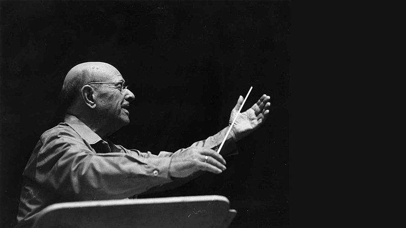 Fotograf�a en blanco y negro del m�sico Pau Casals dirigiendo con intensidad, sosteniendo una batuta y gesticulando con los brazos abiertos sobre fondo oscuro.