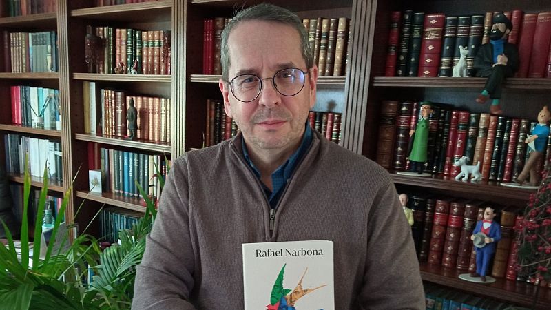 Plano medio de Rafael Narbona con gafas redondas y jersey marr�n, sosteniendo un libro ante una gran estanter�a de madera.