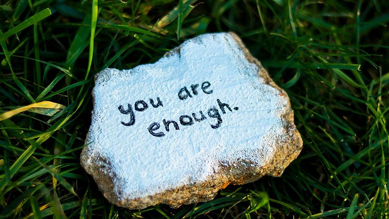 Pequeña piedra pintada de blanco con el mensaje motivador en inglés 'you are enough', apoyada sobre hierba verde.