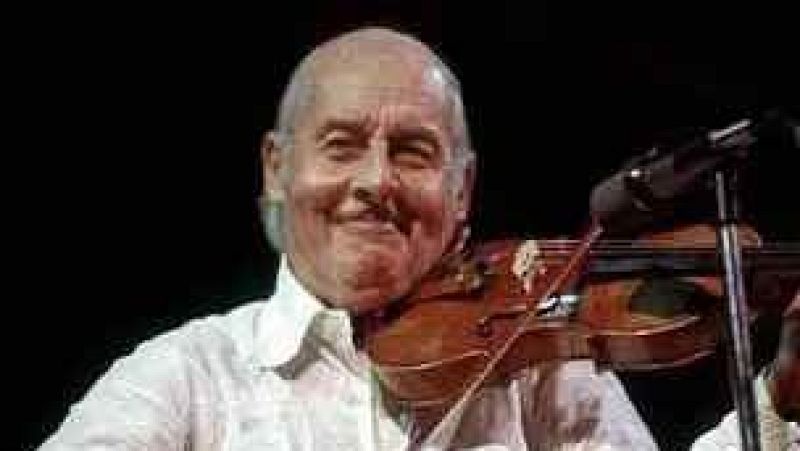 Plano medio del violinista St�phane Grappelli sonriendo con su viol�n ac�stico sobre un fondo oscuro de escenario.