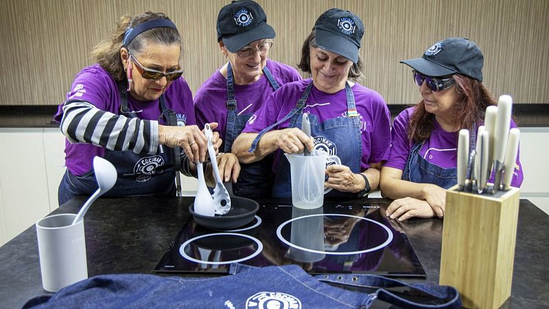 Cuatro mujeres participan en un taller de cocina adaptada alrededor de una isla moderna, realizando tareas culinarias con utensilios y delantales vaqueros.