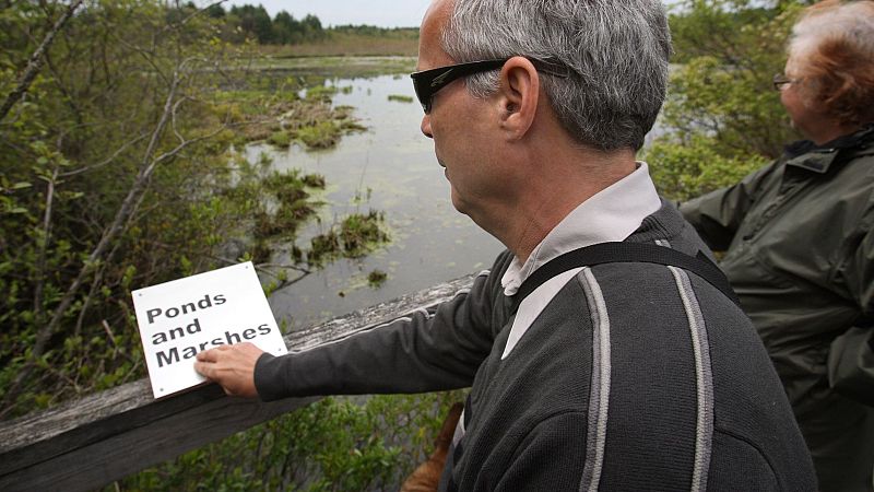 Hombre con discapacidad visual recorre un sendero natural adaptado tocando una placa táctil en la barandilla, con un paisaje de humedal al fondo.